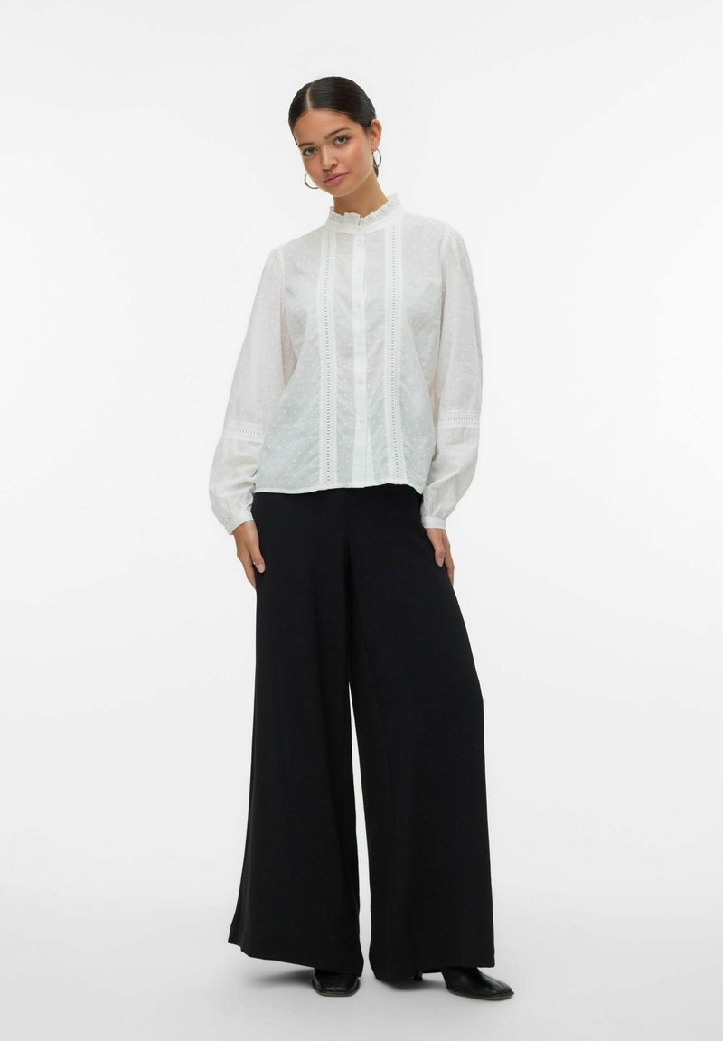Witte blouse met gestructureerde stippen, gerimpelde kraag en verticale plooidetails, gecombineerd met brede zwarte broeken en zwarte schoenen.