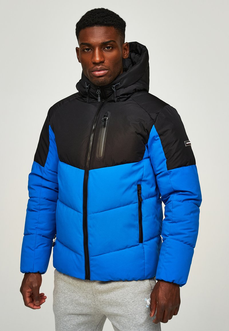 Gesteppten Jacke mit einer schwarzen Kapuze und einem oberen Bereich, der einen blauen unteren Abschnitt hat. Enthält Reißverschlüsse und Logo-Akzente.