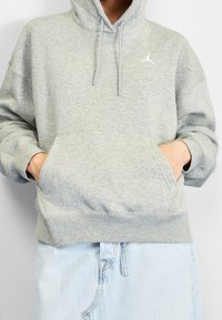 Sudadera con capucha gris con un bolsillo frontal, que presenta un logo blanco. La tela es suave y texturizada, con un ajuste relajado y de gran tamaño.