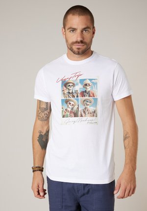 Mann mit kurzen Haaren und Tattoos, der ein weißes T-Shirt mit vier Skeletten in Hüten und Sonnenbrillen und dem Text "Vegas Trip Going Nowhere" trägt.