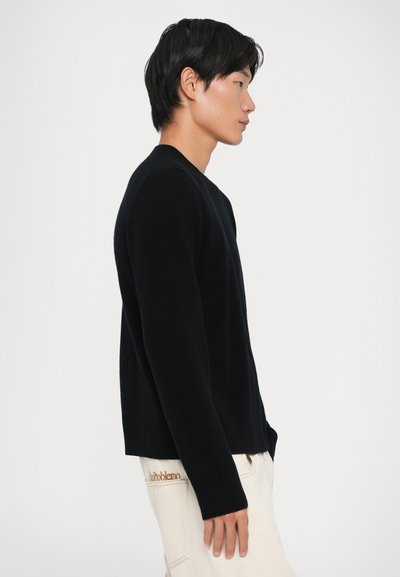 Maglione nero a maglia con maniche lunghe, collo rotondo e orlo a costine; indossato con pantaloni color crema con dettagli ricamati sul lato.