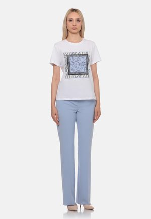 Donna in piedi che indossa una t-shirt bianca con un quadrato decorato e pantaloni azzurri a gamba dritta con tacchi beige.