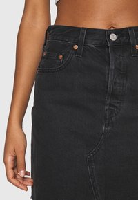 Svart denimkjol med hög midja, kopparfärgade knappar och sidfickor. Tyget har en strukturerad yta med synliga sömmar.