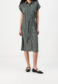 Abito verde con bottoni e maniche corte, con una stampa leopardata in bianco e nero. Cintura in vita e orlo fino al ginocchio. Scarpe nere basse.