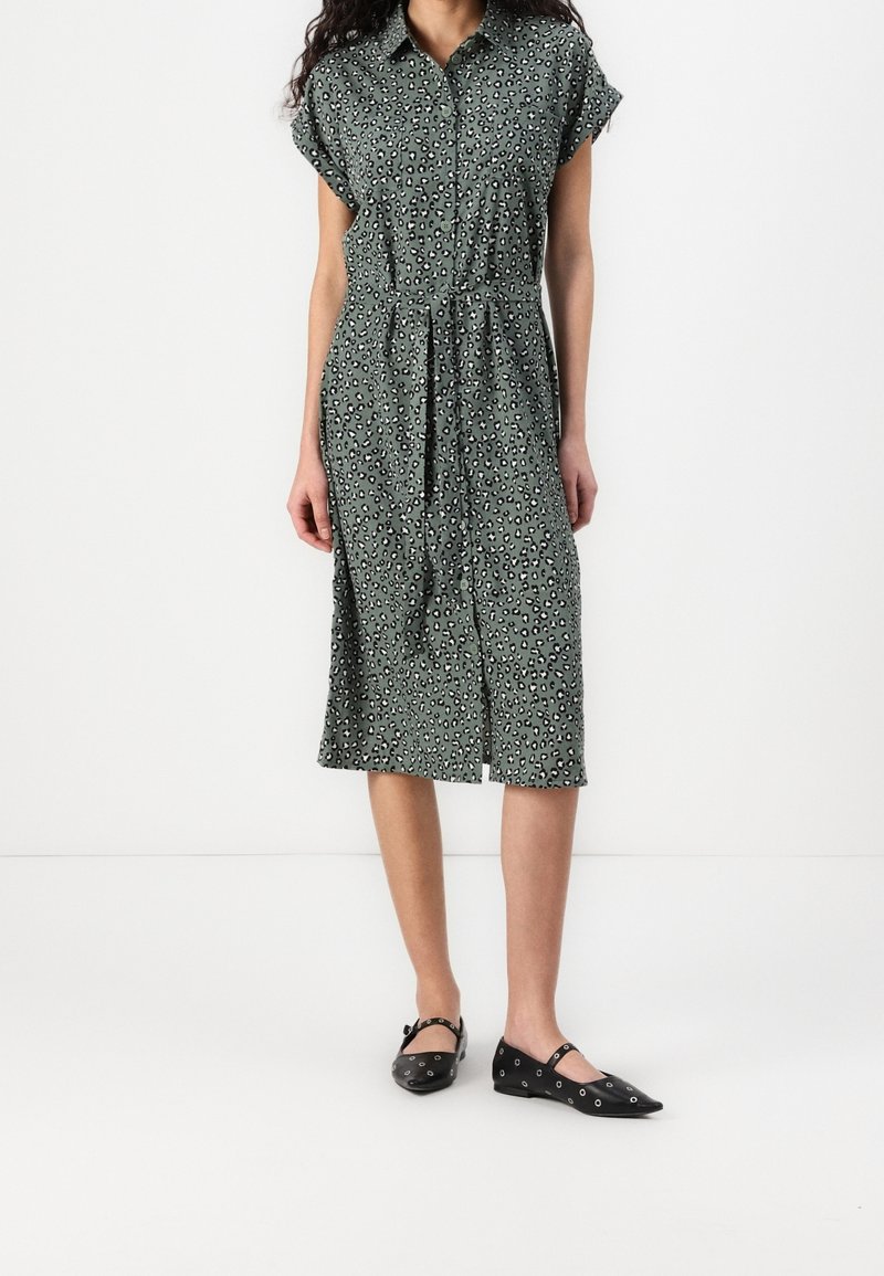 Abito verde con bottoni e maniche corte, con una stampa leopardata in bianco e nero. Cintura in vita e orlo fino al ginocchio. Scarpe nere basse.