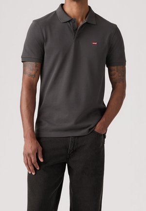 Mężczyzna ubrany w ciemnoszarą koszulkę polo z krótkim rękawem z czerwonym logo Levi's na piersi i czarne spodnie, jedna ręka w kieszeni.