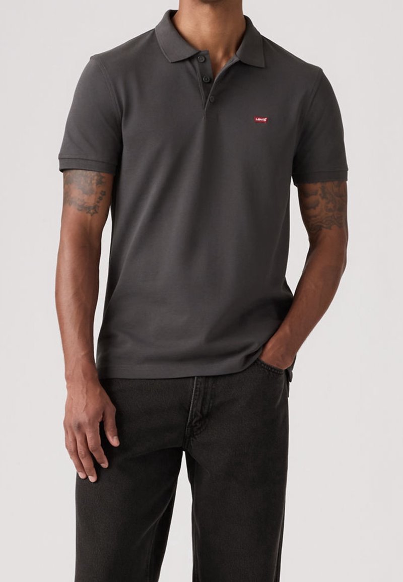 Homme portant un polo gris foncé à manches courtes avec un logo rouge Levi's sur la poitrine et un pantalon noir, une main dans la poche.