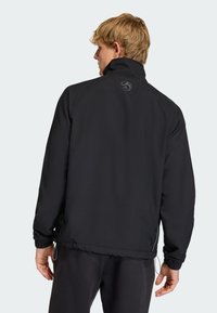 Μαύρο μπουφάν με ψηλό γιακά, ελαστικές μανσέτες και ρυθμιζόμενο τελείωμα με κορδόνι. Διαθέτει λογότυπο στην πλάτη και λεία, ματ υφή.