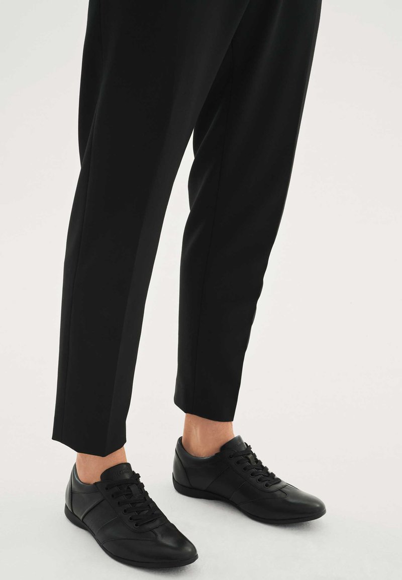 Čierne kožené tenisky so šnurovaním vpredu, s hladkou textúrou a jemným panelovým detailom, v kombinácii s priliehavými čiernymi nohavicami.