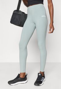 Svetlozeleni legingi z gladko teksturo, z belim logotipom na kolku. V kombinaciji z črnimi športnimi čevlji in črno telovadno torbo.