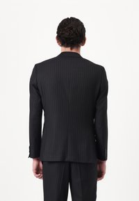 Completo nero a righe sottili con design aderente, caratterizzato da rever a punta, apertura singola e una leggera lucentezza del tessuto, visto da dietro.