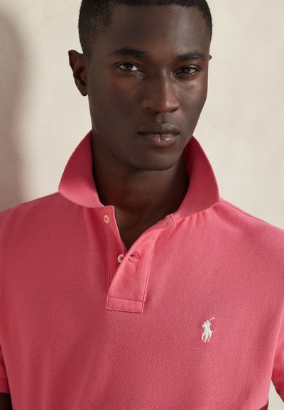 Giovane uomo che indossa una polo rosa con colletto rialzato, con un logo bianco ricamato sul petto sinistro.