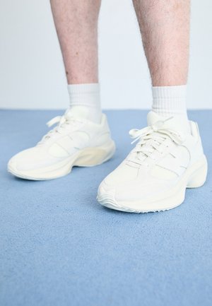 Baskets de sport blanches avec des détails en mesh et en daim, semelle texturée, forme arrondie et fermeture à lacets, portées avec des chaussettes blanches mi-mollet.