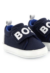 Mornarsko platneni tenisice s bijelom čičak trakom označenom "BOSS." Imaju plavu oblogu i bijeli gumeni potplat. Dizajn s okruglim vrhom.