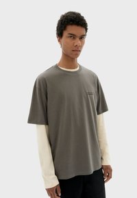 Graues Baumwoll-T-Shirt mit Rundhalsausschnitt, lockerer Passform und kurzen Ärmeln, über einem weißen Langarmshirt getragen. Minimaler Text.