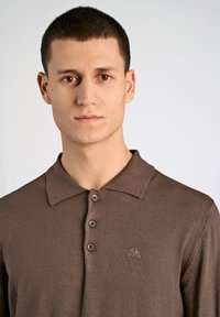 Ruskoppert neulo-paita kauluksella, jossa on kolme nappia ja pieni brodeerattu logo vasemmassa rinnassa. Rakenteellinen kangas ja muotolöydintyylinen design.