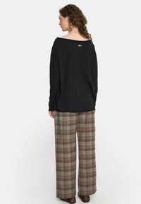 Kobieta z kręconymi włosami nosi luźną czarną bluzkę z długim rękawem i brązowe, w kratę, szerokie spodnie z wysokim stanem, stojąc na tle jednolitym.