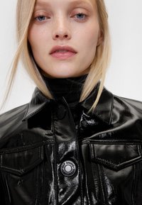 Blizgi juoda patentuota džinsinė striukė su smailiu apykakliu, priekinėmis kišenėmis ir didelėmis sagų detalėmis. Lygi tekstūra su prigludusiu dizainu.