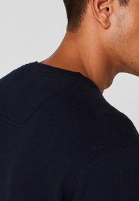 Pull en tricot bleu marine avec un col rond, montrant un gros plan de la couture de l'épaule et du tissu texturé. Fini lisse et doux.