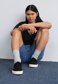 Black sneakers with white soles, gray ribbed socks, denim shorts, and a black t-shirt with a logo, sitting against a light wall.