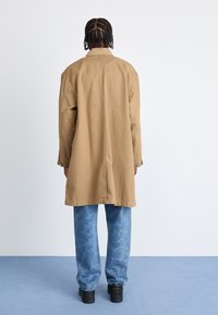 Tan lang frakke med en afslappet pasform, vist over blå mønstrede jeans. Jakken har en krave og knapdetaljer på ærmerne.