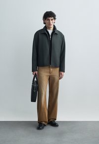 Zwarte ritsjack over een wit shirt, gecombineerd met wijde, lichtbruine broekspijpen. Zwarte schoenen en een zwarte tas maken de outfit compleet.