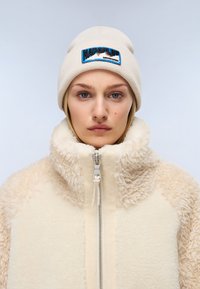 Giacca in pile beige con colletto alto e chiusura con zip, con texture morbida sulle maniche. Berretto in maglia beige con logo blu ricamato.