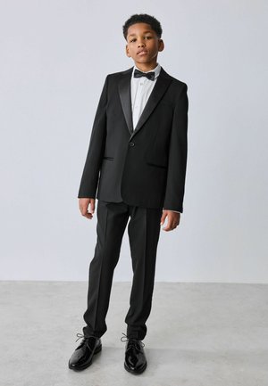 Poiss, kes kannab musta fraaki, valget särki, musta kikilipsu ja läikivaid musti kingi, seisab neutraalse tausta ees.