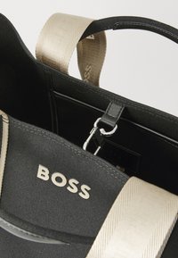 Borsa nera in tela con manici beige e dettagli in metallo argentato. Presenta un manico superiore, il logo "BOSS" in evidenza e una tasca interna con zip.