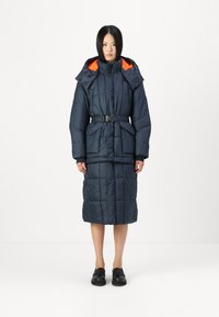 Cappotto imbottito blu navy con cappuccio foderato arancione, vita con cintura e texture trapuntata. Presenta tasche frontali e lunghezza fino a metà polpaccio.