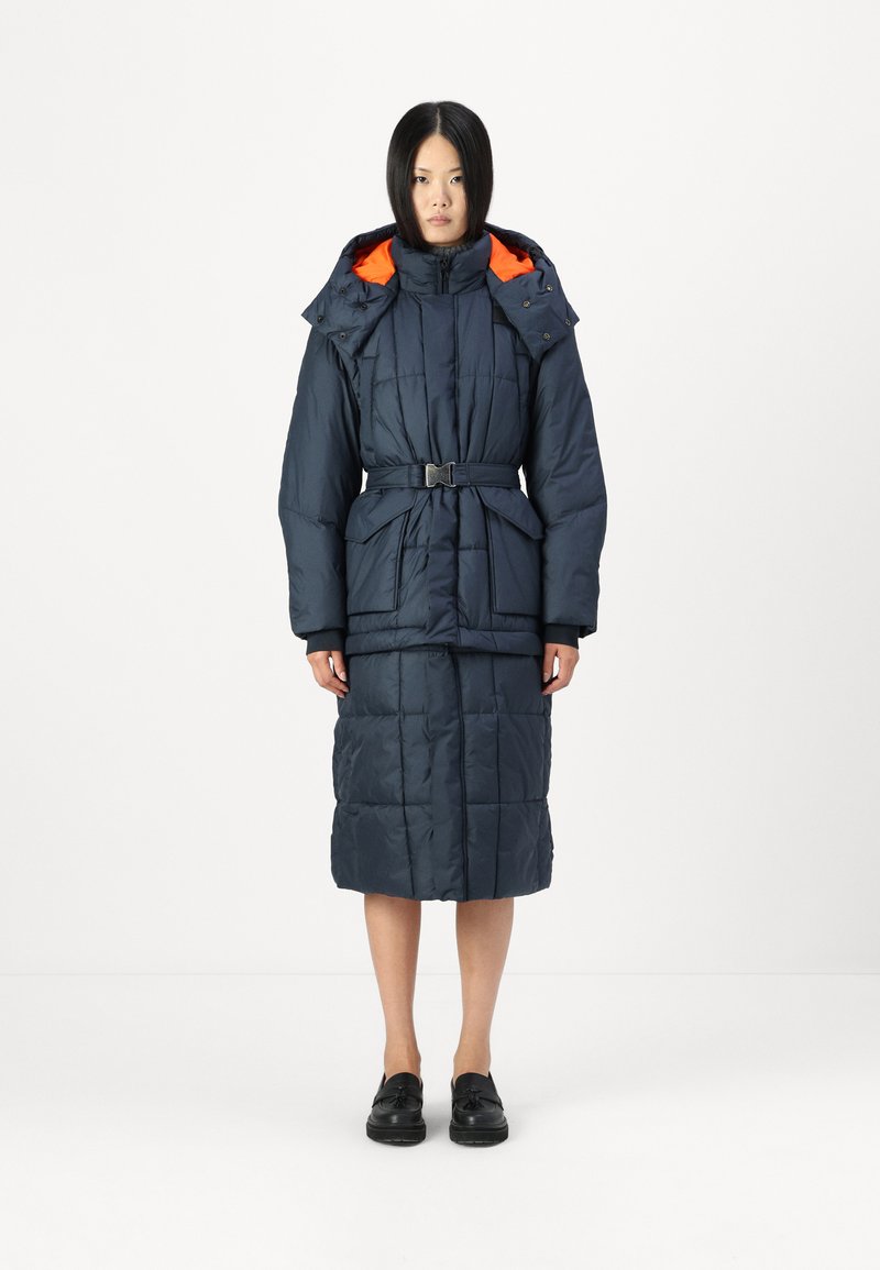 Cappotto imbottito blu navy con cappuccio foderato arancione, vita con cintura e texture trapuntata. Presenta tasche frontali e lunghezza fino a metà polpaccio.