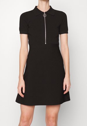 Kvinde iført en sort kortærmet kjole med krave og lynlås foran, stående mod en ensartet lys baggrund.