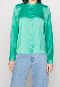 Camicia lunga verde in satin con colletto, dotata di chiusura con bottoni frontale e una superficie liscia, abbinata a jeans azzurri chiari.