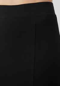 Gros plan d'une personne portant un pantalon noir taille haute avec une large ceinture élastique et un détail de couture verticale.