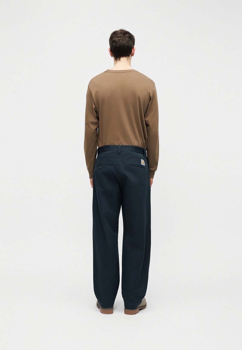 Mann steht mit dem Rücken zur Kamera, trägt ein braunes Langarmhemd, dunkle marineblaue weite Hosen mit Gesäßtaschen und hellbraune Schuhe vor einem einfarbigen Hintergrund.