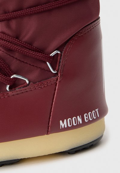 Prim-plan al unei Moon Boot în nuanță de bordeaux, cu textură din material textil și piele, cu ochiuri din metal, șireturi asortate și talpă bej vizibilă.