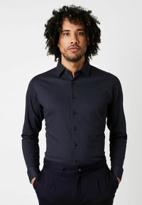 Navyblaues Langarm-Hemd mit Knöpfen, das ein kleines Punktmuster aufweist, slim fit und einen spitzen Kragen hat. Getragen mit dunklen Hosen, Hände in den Taschen.