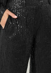 Zwarte paillettenbroek met verticale strepen van glanzende pailletten, voorzien van zijzakken en een soepele textuur, die het licht vangt met reflecterende details.