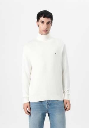 Hvit turtleneck genser laget av mykt strikket stoff, med lange ermer og ribbestrikkede mansjetter, med en liten logo på brystet.