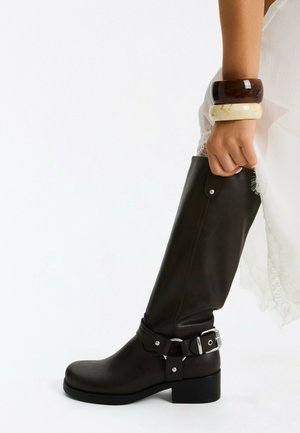 Main tenant une botte noire montante avec boucle argentée, portant un vêtement en dentelle blanche et deux larges bracelets, un brun foncé et un crème.