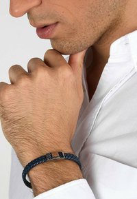 Navy braided leather bracelet with silver-toned accents and a metallic emblem, set against a white shirt.