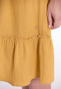 Senfgelbe Kleid aus einem strukturierten, gekräuselten Stoff. Mit einem Rüschensaum und einem kleinen runden Etikett auf der Vorderseite. Maße und Details nicht sichtbar.