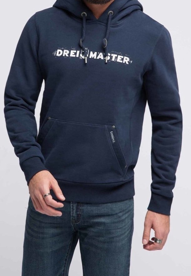 Homme portant un sweat à capuche bleu marine avec le texte "DREIMASTER", une poche avant et des cordons, associé à un jean bleu et des bagues argentées aux deux mains.