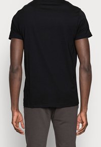 Hombre con una camiseta de manga corta negra lisa y pantalones oscuros, de pie de espaldas con los brazos relajados a los lados frente a un fondo liso.