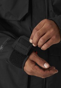 Chaqueta impermeable negra con ajuste en los puños mediante velcro. Primer plano de manos ajustando el puño, mostrando texturas contrastantes y detalles de diseño sin costuras.