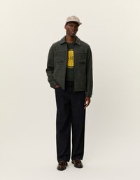 Tumši zaļa teksturēta jaka ar divām krūšu kabatām, vilkta pāri melnai grafiskai T-kreklam. Apvienota ar tumšām biksēm un brūnām kurpēm ar auklēm.