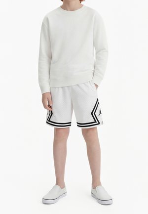 Personne portant un sweat-shirt blanc à manches longues, un short de basket blanc avec des bordures noires et des chaussures blanches sans lacets, debout avec une main dans la poche.