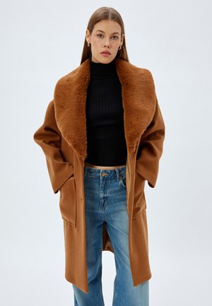 Abrigo de lana marrón con un gran y suave cuello de piel sintética, de frente abierto y con bolsillos laterales; usado sobre un cuello de tortuga negro de canalé y jeans azules.