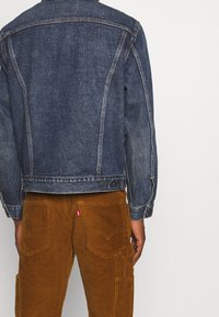 Chaqueta de mezclilla de color azul oscuro con un corte clásico, que cuenta con dos bolsillos frontales y costuras en contraste. Lleva puesta sobre pantalones de pana marrones.