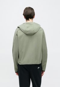 Zielona kurtka z kapturem o luźnym kroju, miękkiej fakturze i detalach szwów, zestawiona z czarnymi sportowymi spodniami z małym logo.